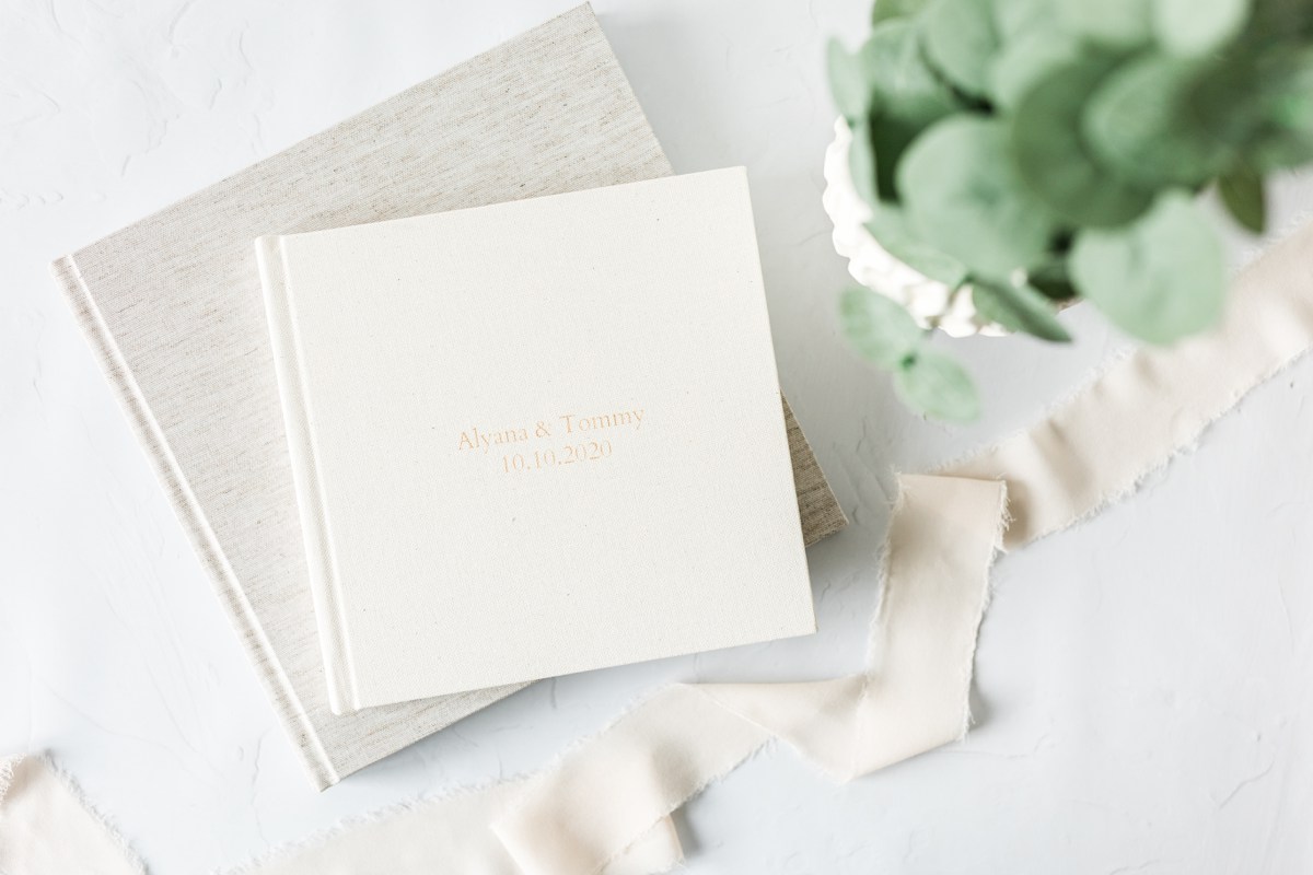 Two wedding albums stacked on top of each other in a flat lay style. This light and airy photography shows a tan album and a cream colored album with a gold inscription of the couple's names and wedding date. Wedding albums are the perfect way to tell the story of a wedding day in vivid detail.