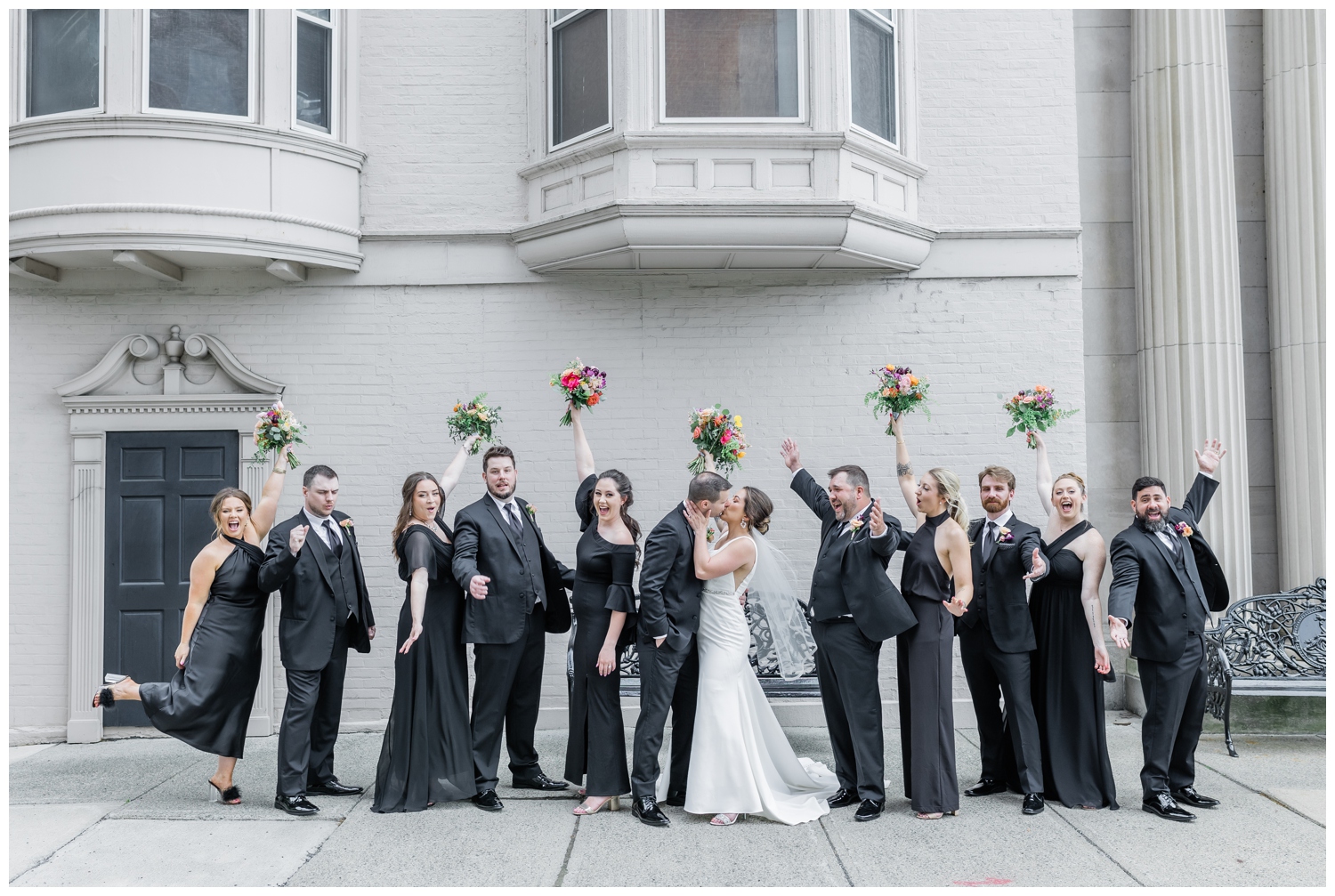 Bridal party all in black with with bright colorful bouquets cheering for the bride and groom as the newlyweds kiss in front go the Franklin Plaza Ballroom in Troy, NY.