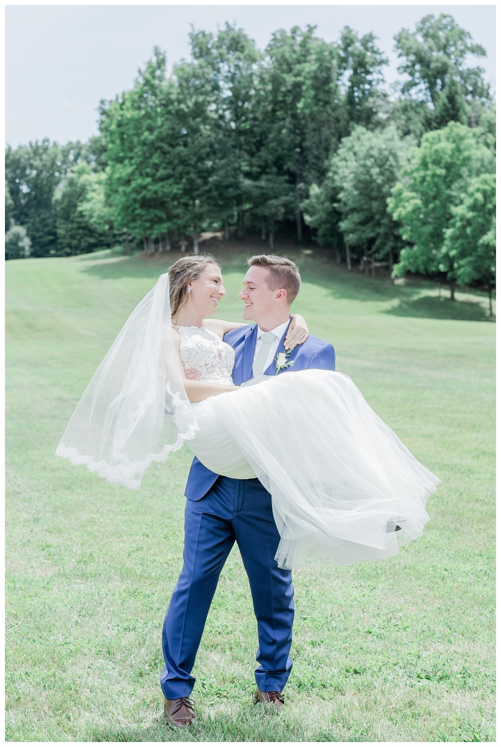 Wedding photo of a couple in a field at Hayloft at the Arch. Groom is wearing a navy blue suit and the bride is wearing a high neckline wedding dress with lots of tulle and a vail.