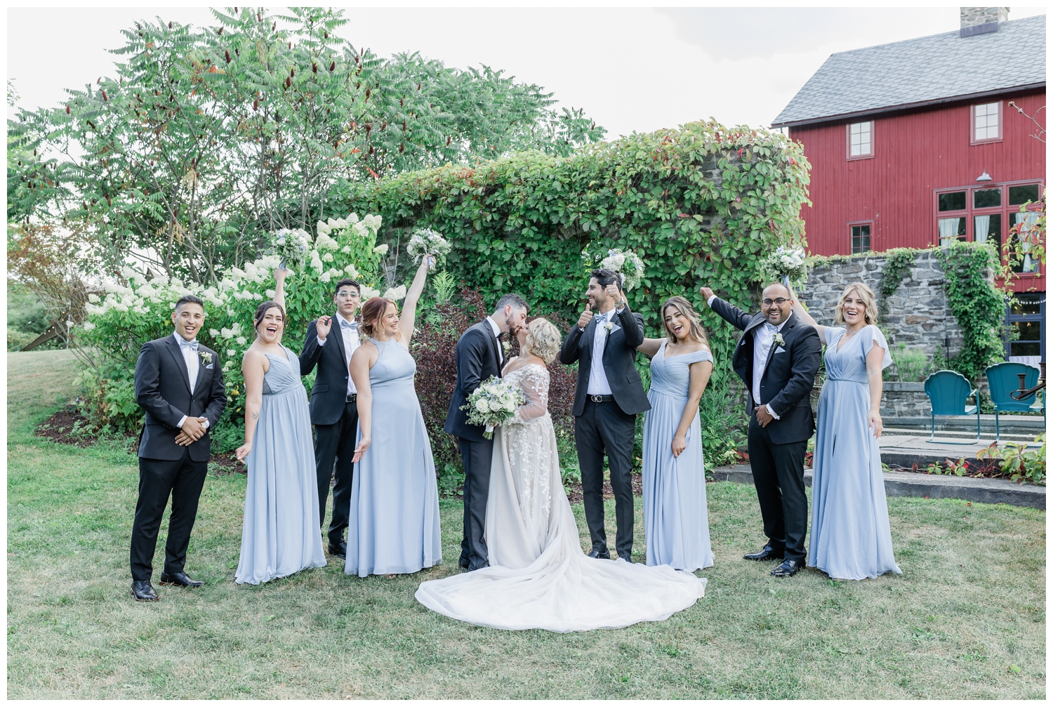 Wedding party cheering for the bride and groom as they kiss. Groomsmen are wearing black suite with a powder blue bowtie while the bridesmaids are in different style dresses but all in the matching light blue color. 