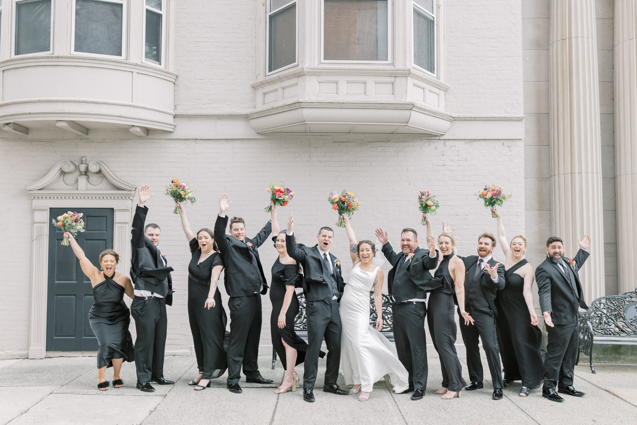 A bridal party at a wedding throwing their hands up in the air in celebration of the Bride and groom who just got married. They are all wearing black dresses and black suits with colorful wedding bouquets. This wedding took place at Franklin Plaza in Troy, NY and was photographed in a light and airy style. 