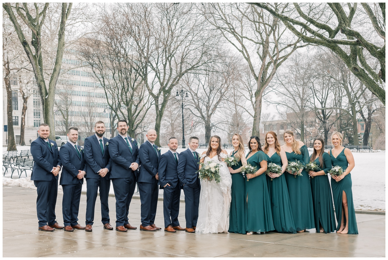 Wedding party on a wedding day all in a row. Bridesmaids are wearing beautiful deep green dresses while the groomsmen are in navy blue suits with matching green ties. This wedding was in downtown Albany, NY by the Capital building on a snowy March day.