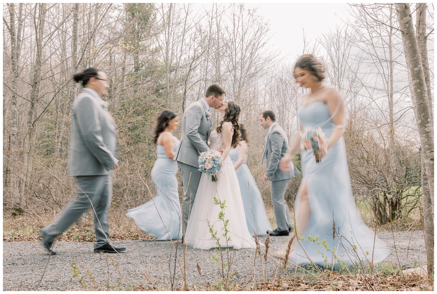 Bride and groom kissing as their wedding party surrounds them in gray suits and light blue dresses. 
