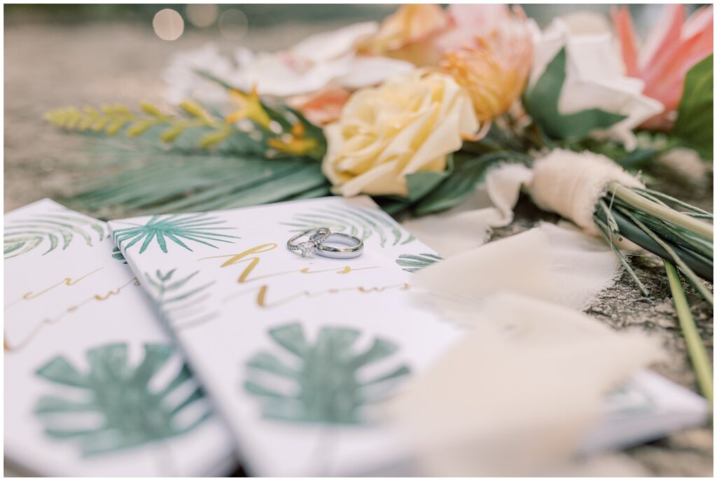 Tropical detail shot of wedding rings on vow books with a colorful bouquet. 
