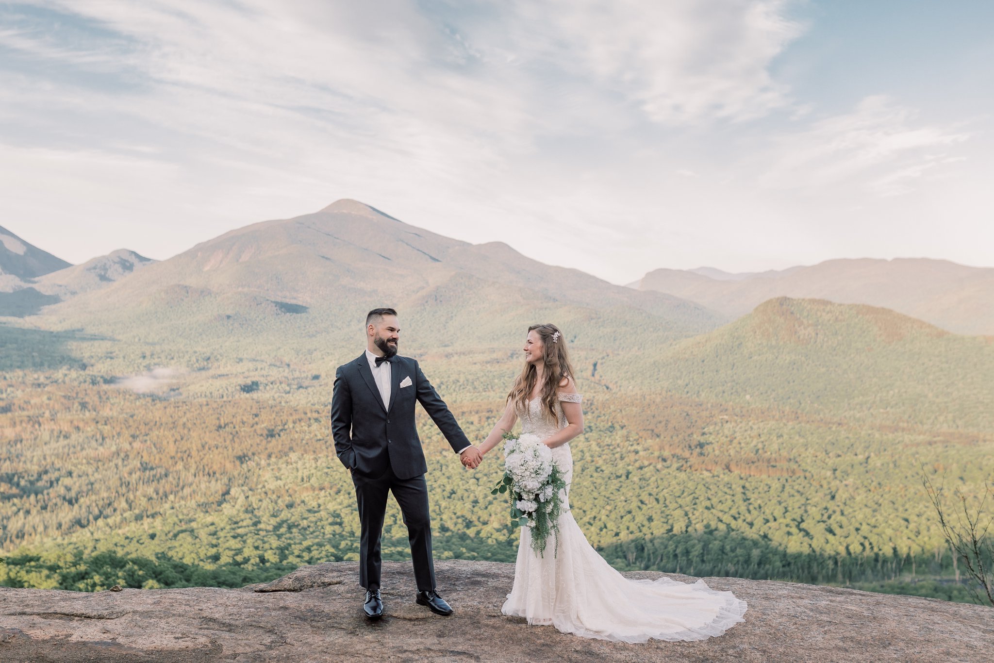 Bride and groom holding hands on the summit of a mountain with Adirondack mountains in the background.