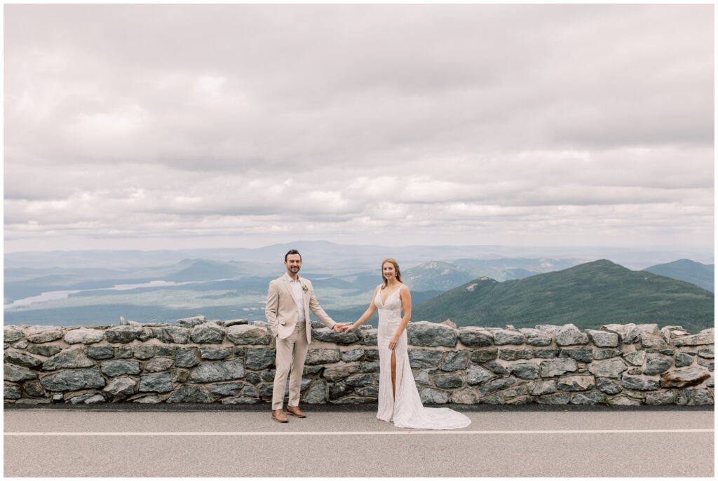 Bride and Groom holding hands by a rock wall with the Adirondack mountains in the background. 
