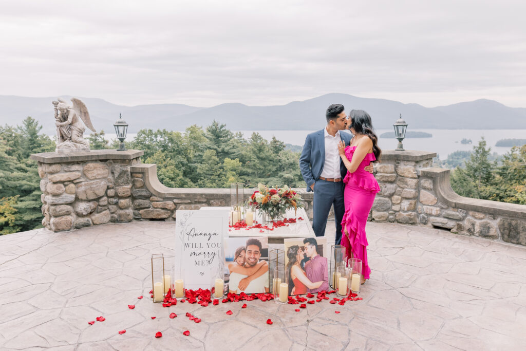 Couple kissing in front of a sign saying "Will you marry me?" at a castle over looking Lake George in New York.