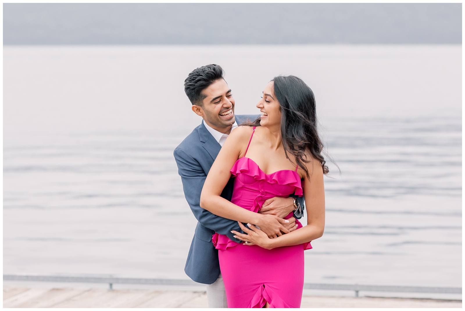 Man hugging is partner from behind as they laugh together standing in front of a lake.
