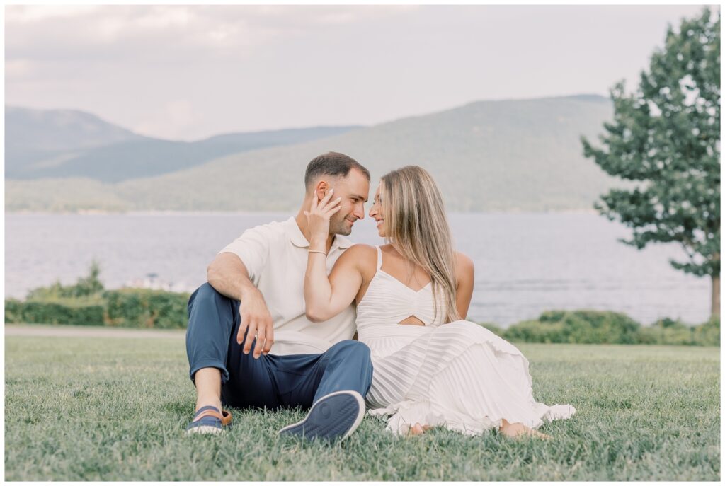 Couple sitting on the ground in front of the lake getting engagement photos taken during their Lake George Engagement Session.
