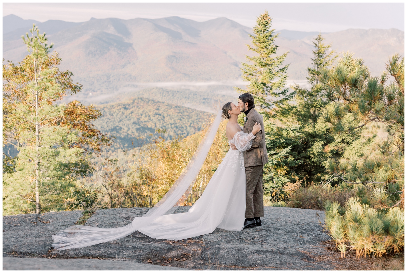 Wedding couple kissing while standing on a rock with mountains in the background.
