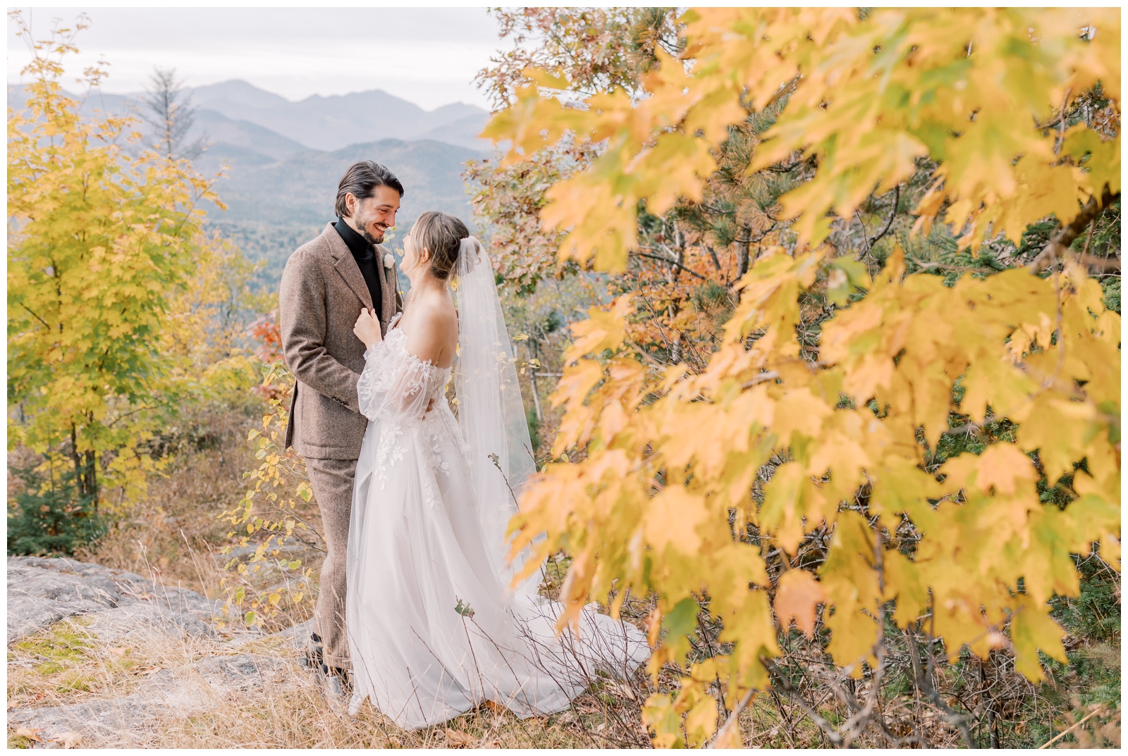 Husband and Wife smiling at each other standing next to a yellow tree with a mountain range in the background.