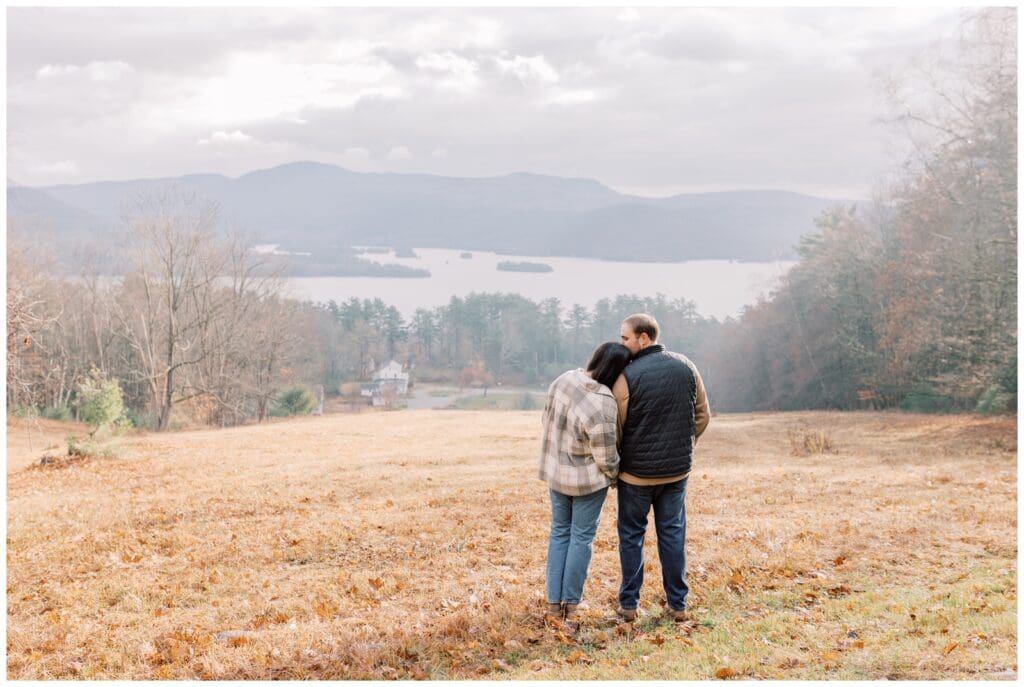 Couple standing in a field overlooking a lake in the Adirondacks after their proposal in the Fall.
