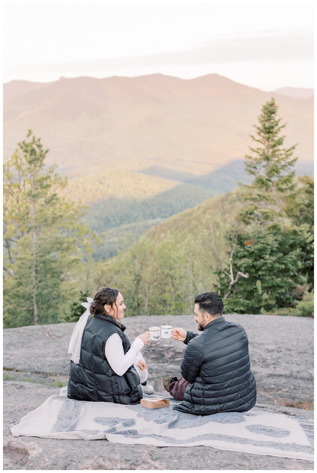 Couple sitting on a blanket on top of an Adirondack mountain at sunrise drinking coffee together.
