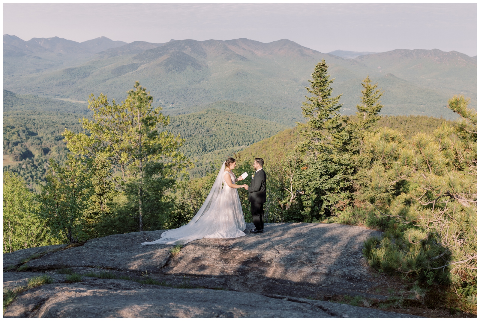Bride and groom standing at the peak of a mountain overlooking a mountainous view while reading their vows during their elopement ceremony at sunrise.
